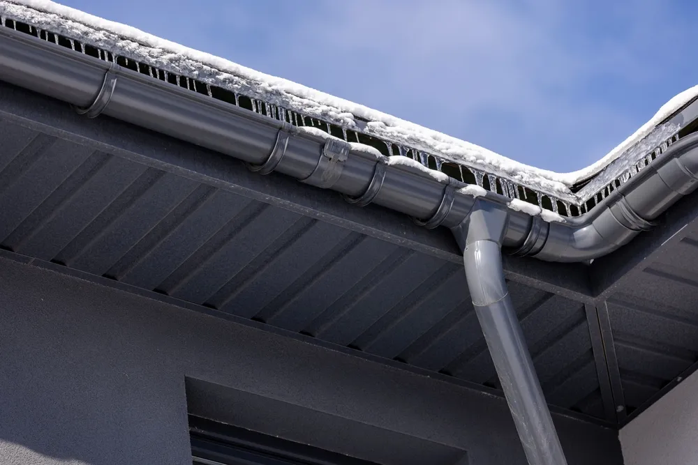 Modern metal gutter system with icicles and snow-covered eaves, highlighting winter conditions and roof drainage against a clear blue sky.